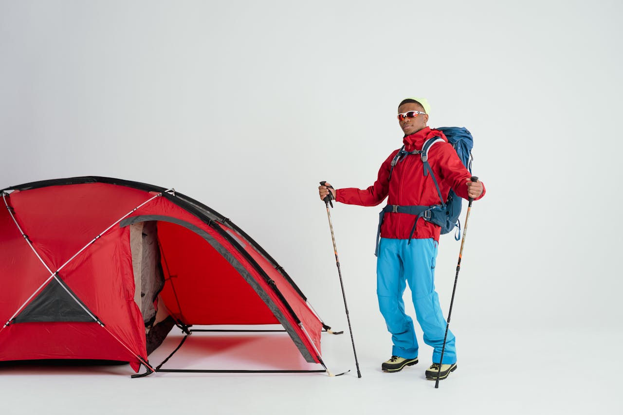 A man dressed for winter camping stands by a red tent with trekking poles.